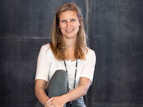 Porträt Bild Carmen Gründerin von Soulbase. Sie trägt ein weisses T-Shirt und einen Türkise Yogahose zudem eine Malakette im Yogastudio YogArosa