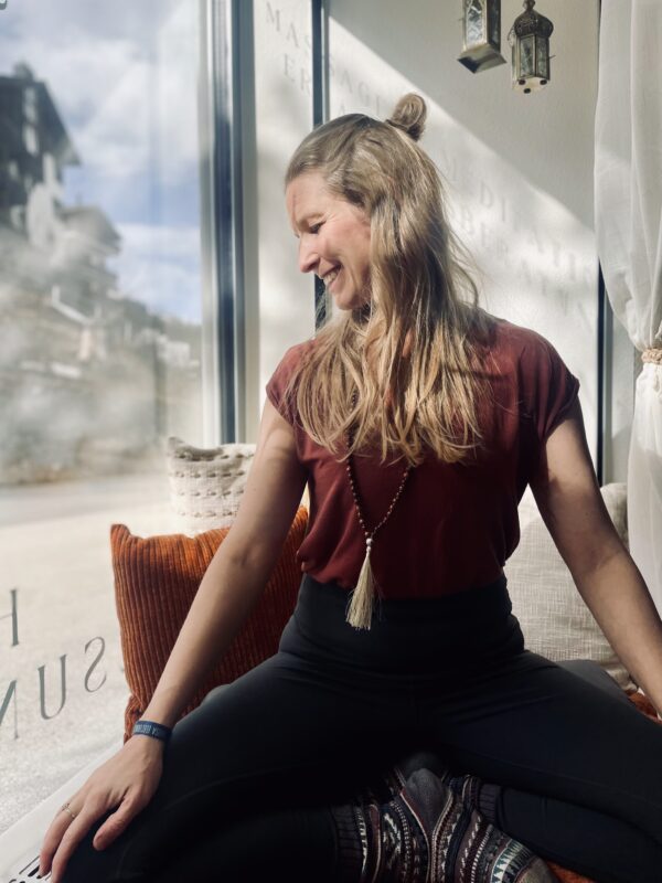 Schneidersitz Meditationspose, lachende Frau schaut zur Seite und hat blondes Haar und trägt ein rostrotes Shirt
