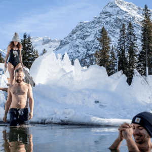 Eisbadi Arosa, die erste Eisbadi in der Schweiz. 3 Freunde die dort bei schönen Wetter und Bergkulisse umgeben von Eisplatten baden gehen. Es sind eine Frau und zwei Männer mit Badekleidung und Mützen.