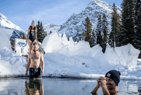 Eisbadi Arosa, die erste Eisbadi in der Schweiz. 3 Freunde die dort bei schönen Wetter und Bergkulisse umgeben von Eisplatten baden gehen. Es sind eine Frau und zwei Männer mit Badekleidung und Mützen.