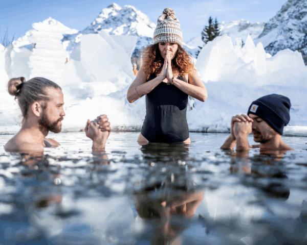 Eisbadi Arosa, zwei Männer und eine Frau sind im Eiswasser. Die Frau trägt eine Mütze und einen schwarzen Badeanzug und hält die Hände zur Namaste Pose. Der See ist zugefroren und Eisplatten sind dekorativ am Rand aufgestellt. Untersee Arosa Graubünden