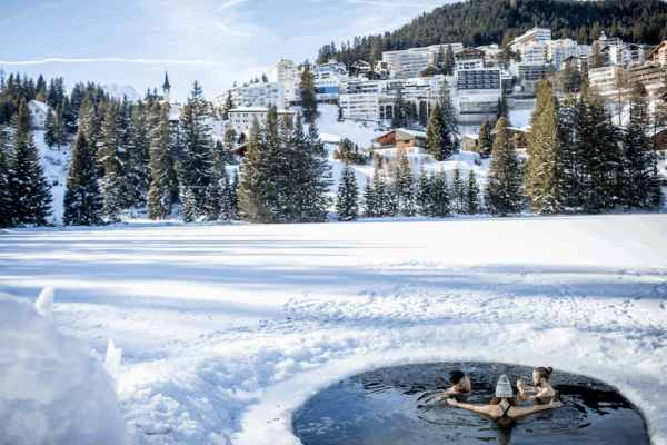 Eisbadi Arosa, Drohnenbild mit Blick auf den zugefrorenen Untersee und Arosa, bei schönen Wetter. 3 Leute sitzen in schönen Ambiente im Eisloch