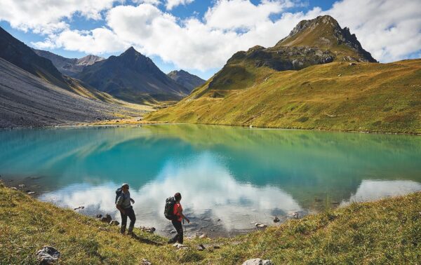 Schwellisee in Arosa. Zwei Wanderer umrunden den See bei guten Wetter. Die Aufnahme bietet Blick ins Tal, die Wiesen sind grün und der See türkis