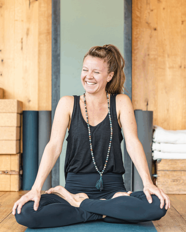 Portrai von Ronja Gründerin von Soulbase. Sie sitzt im Schneidersitz trägt ein schwarzes T Shirt und eine schwarze leggins sowie eine selber gemachte Malakette. Das Foto ist bei Yogarosa im Yogastudio entstanden
