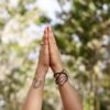 2 Hände in Grossaufnahme in Namaste Haltung. Die Frau trägt Armeinreifen udn ein Tattoo, das ganze ist in freier Natur.